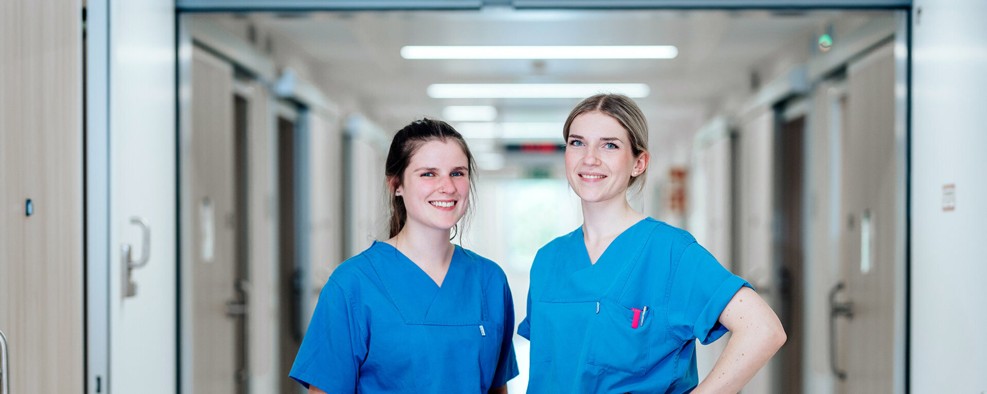 Zwei lächelnde Pflegefachkräfte, im Hintergrund der Krankenhausflur